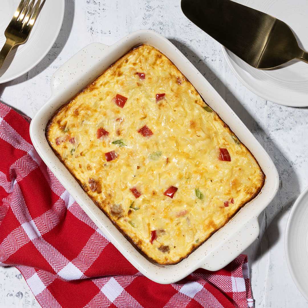 A top-down close-up view of a golden-brown, cheesy hashbrown casserole in a white, speckled baking dish. The casserole is garnished with chopped red and green bell peppers.