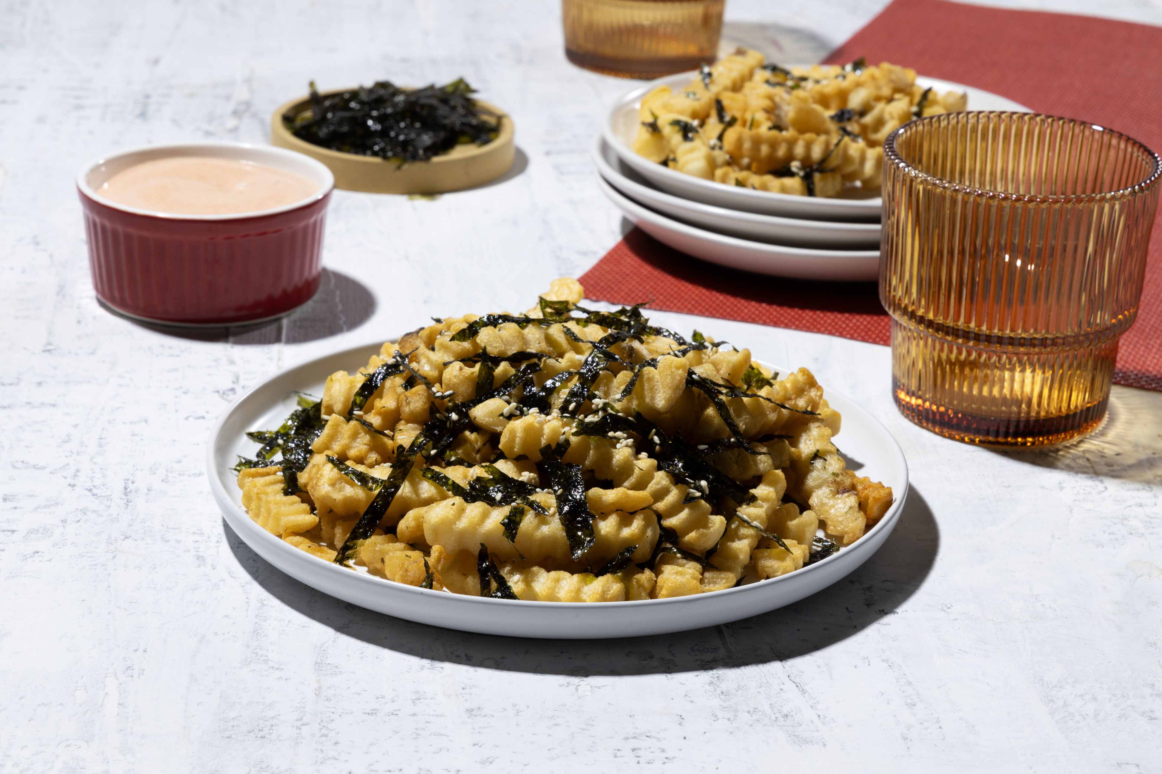 A close-up view of a plate piled high with golden-brown Ore-Ida crinkle-cut fries. The fries are generously topped with shredded nori seaweed and a sprinkle of sesame seeds. A small, red ramekin filled with a creamy, pinkish Yum Yum sauce sits to the left of the fries, and a stack of two similar plates with fries can be seen slightly out of focus to the right. A blurred, ribbed, amber-colored drinking glass is positioned to the far right of the frame. The background is a textured, off-white surface, and the lighting is bright and even, highlighting the textures and colors of the food.