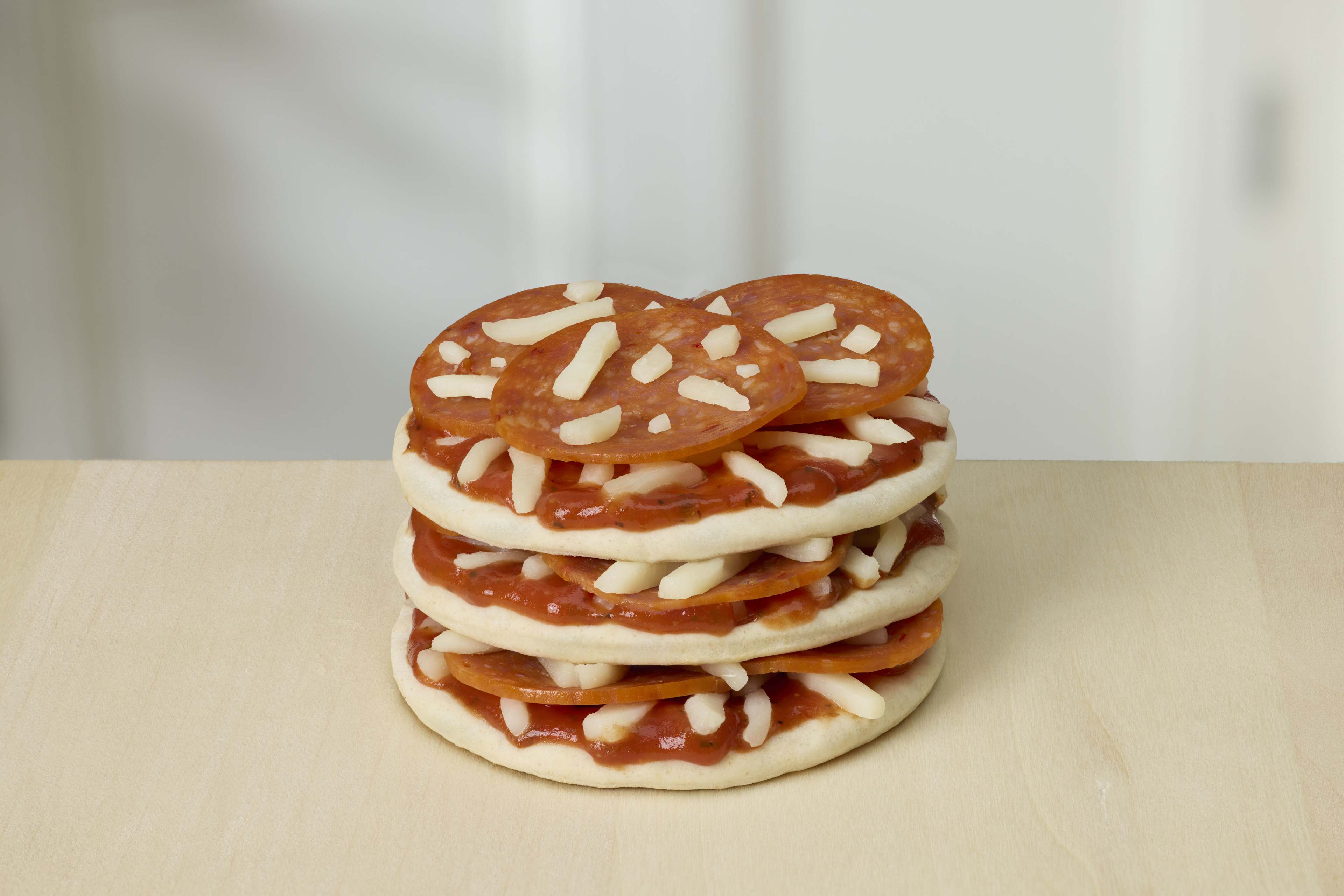 Three cold, unbaked Lunchables pepperoni pizzas stacked on a light wood table, topped with shredded cheese and pepperoni.