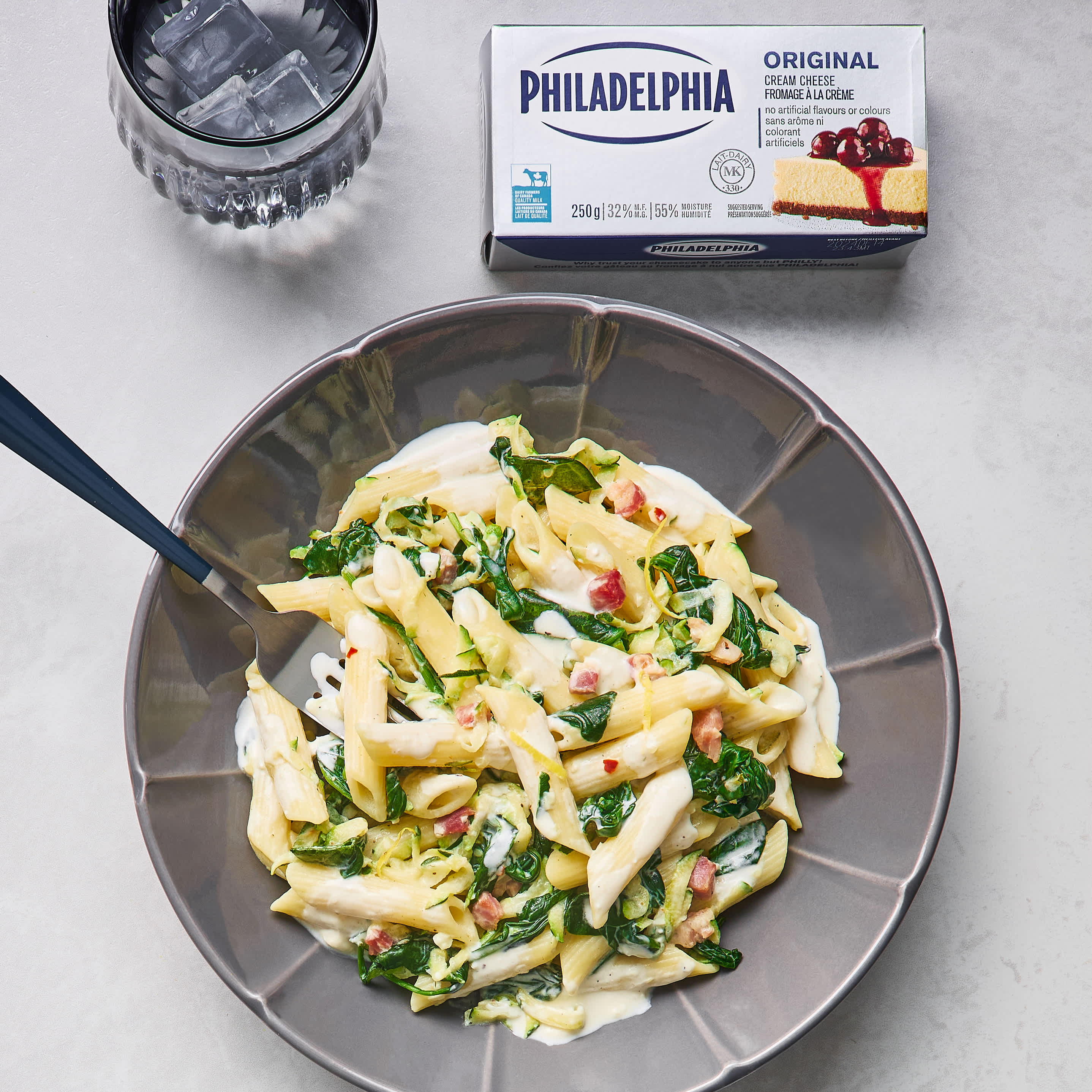 Penne pasta with zucchini, spinach, and lemon sauce in a grey bowl near a Philadelphia Original Cream Cheese box.