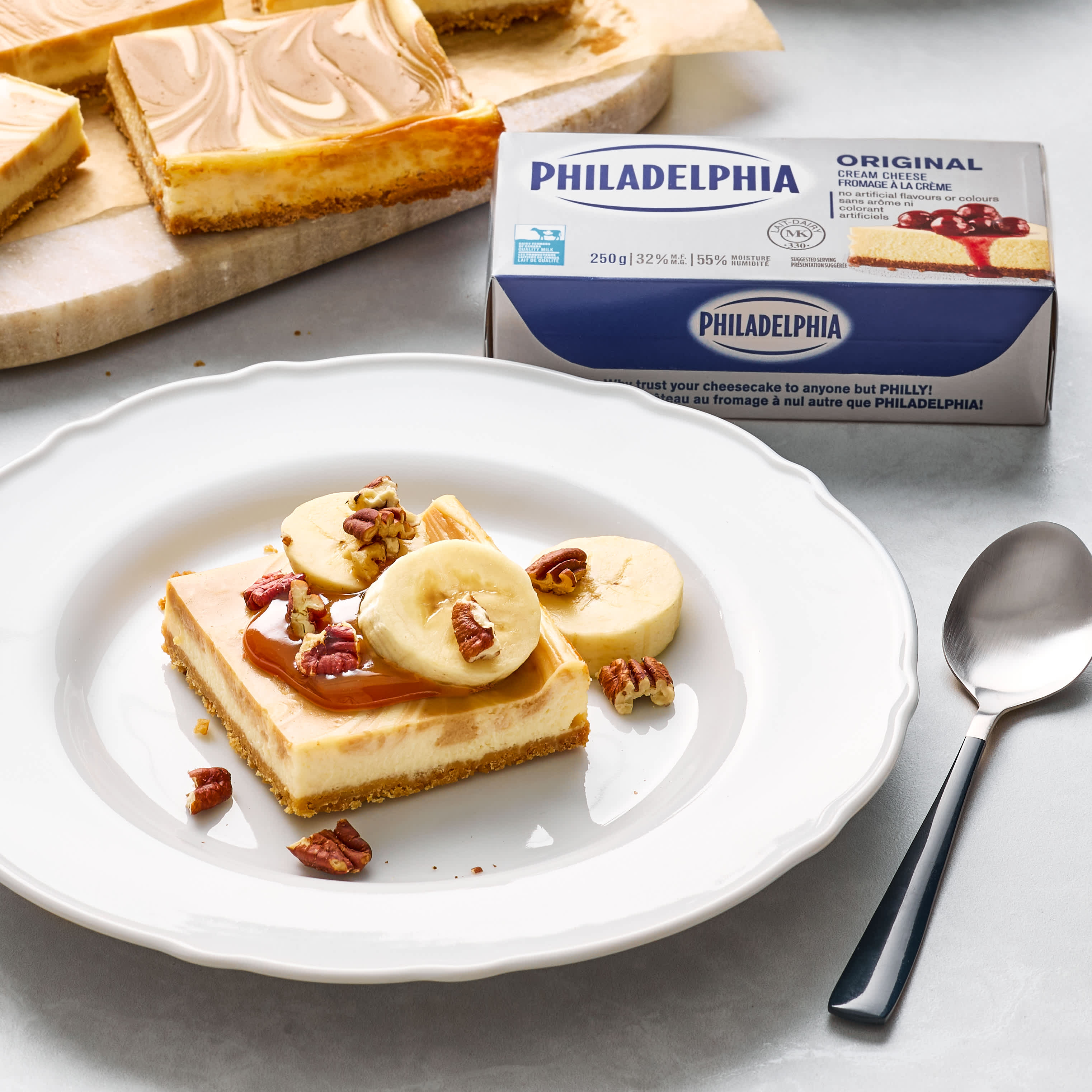 Sheet Pan Dulce de Leche Cheesecake slice with bananas and pecans by a Philadelphia Cream Cheese 250g box.