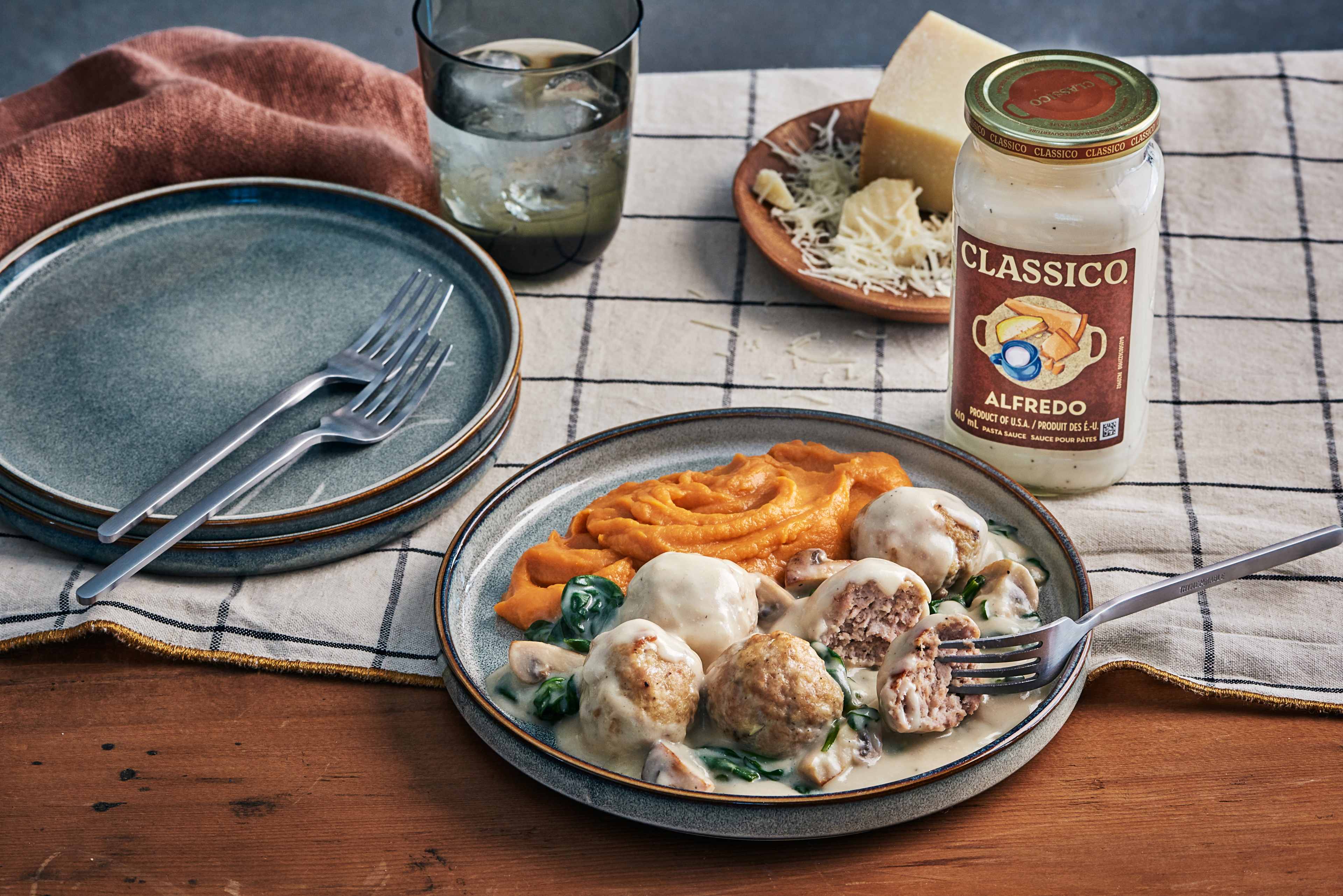 Une assiette de savoureuses boulettes de viande italiennes nappées d'une sauce Alfredo crémeuse, accompagnées d'une portion de purée de patates douces à la couleur orange vif. Un pot de sauce Alfredo Classico se trouve à l'arrière-plan, à côté de parmesan râpé, laissant deviner l'ingrédient principal. Deux assiettes et fourchettes supplémentaires reposent sur une nappe à carreaux, complétant le décor invitant.