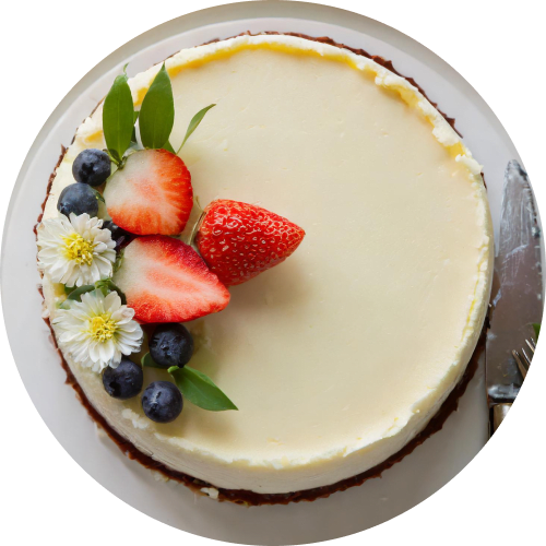 Overhead shot of a whole cheesecake on a white plate. The cheesecake is garnished with fresh strawberries, blueberries, white flowers, and green leaves. A knife is placed next to the cake, ready to serve.