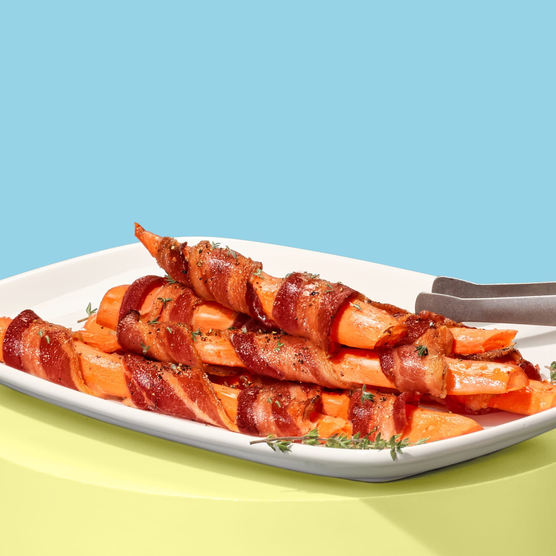 A platter of roasted carrots wrapped in crispy bacon, garnished with thyme on a light blue and yellow background.