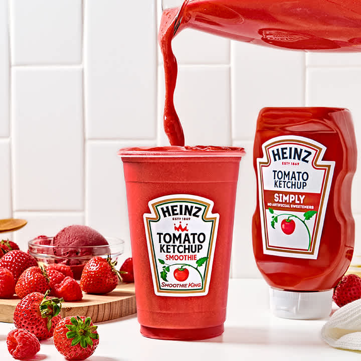 A vibrant red Heinz Tomato Ketchup Smoothie being poured into a clear cup, with a Heinz Simply Tomato Ketchup bottle, fresh strawberries, and raspberries on a white countertop, highlighting a unique and flavorful recipe.