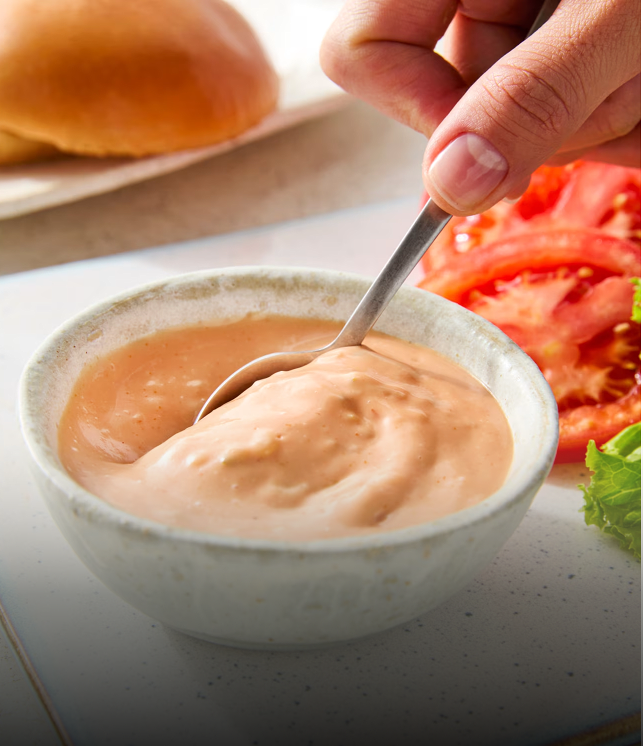 Gros plan d'un bol de sauce crémeuse pour hamburger avec une cuillère, à côté des ingrédients du hamburger.