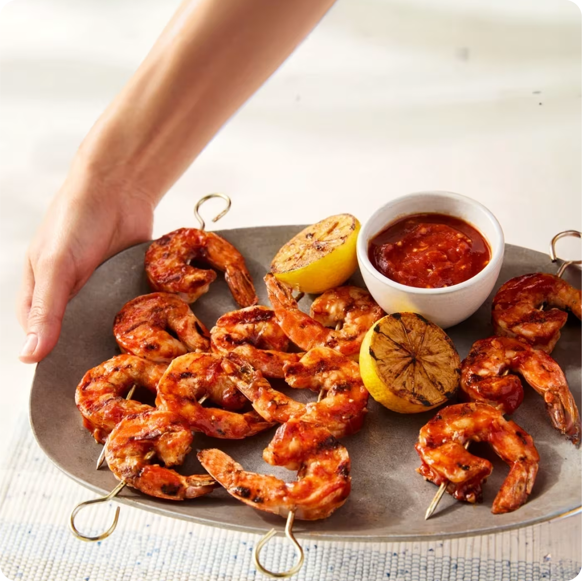 Brochettes de crevettes grillées avec sauce et moitiés de citron grillé sur un plateau argenté tenu par une main.
