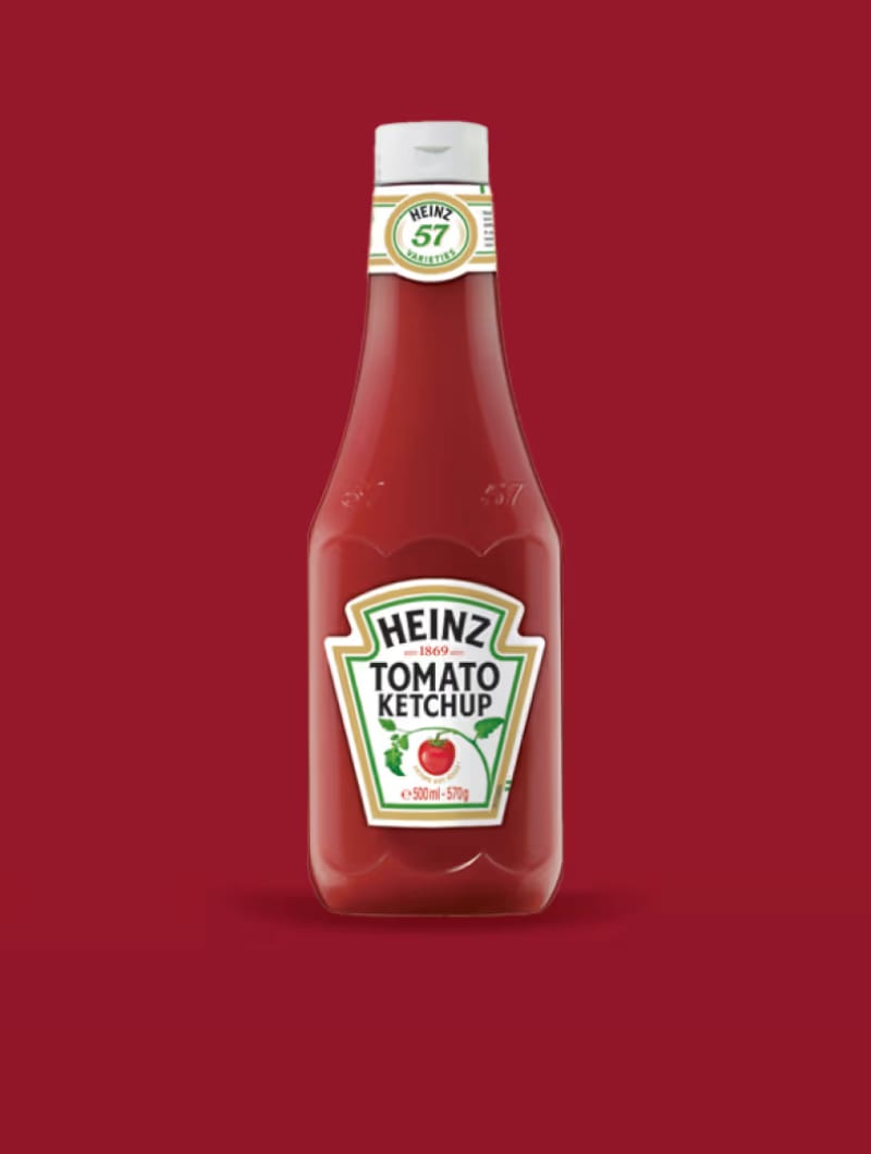 Červená láhev Heinz Ketchup s bílou etiketou a vyobrazením rajčete, na vínově červeném pozadí.