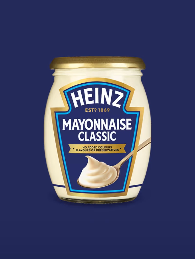 Sklenice Heinz Mayonnaise Classic s bílou etiketou a lžící majonézy na tmavě modrém pozadí.