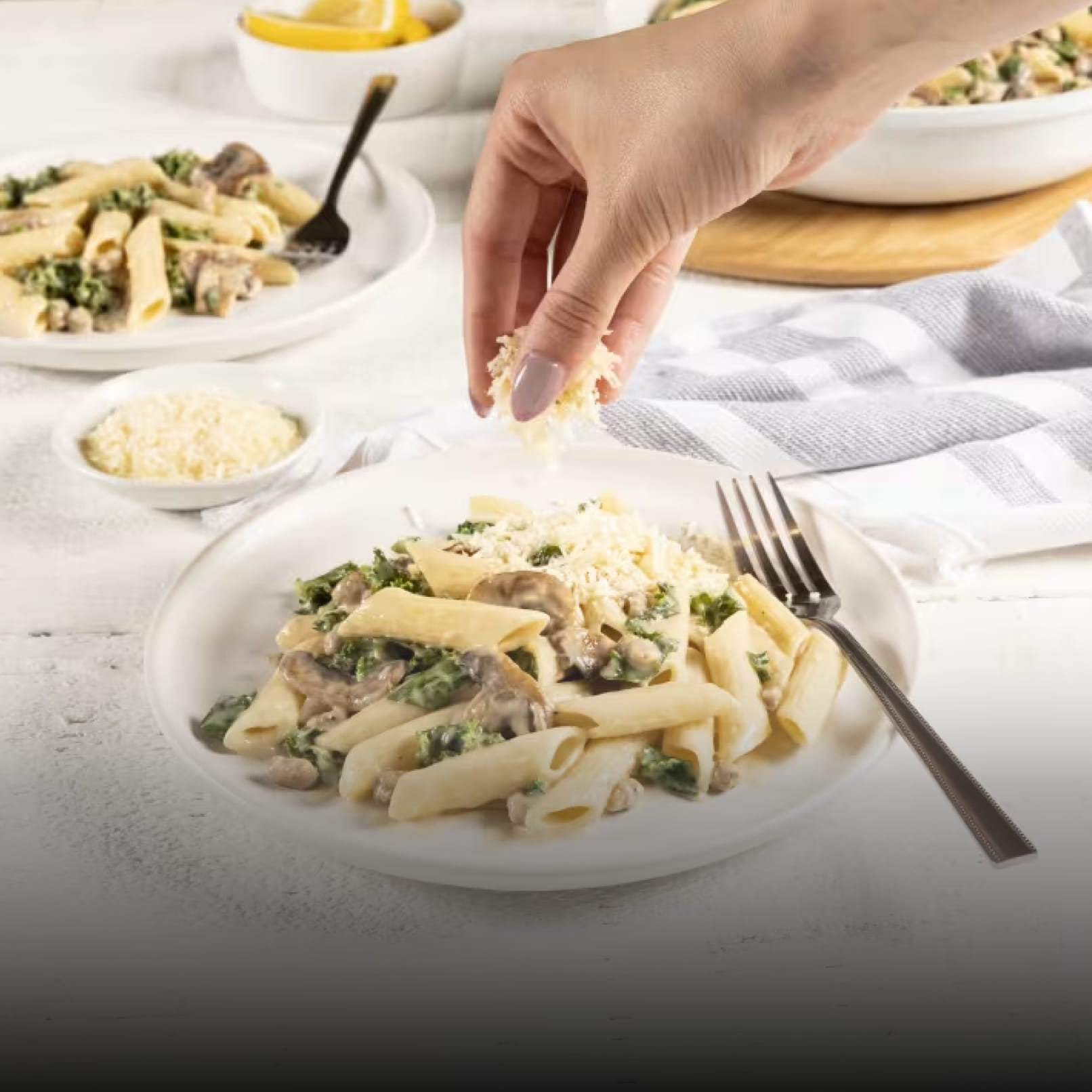 A plate of creamy pasta with sausage, mushrooms, and kale is presented on a white wooden table. A hand sprinkles grated parme