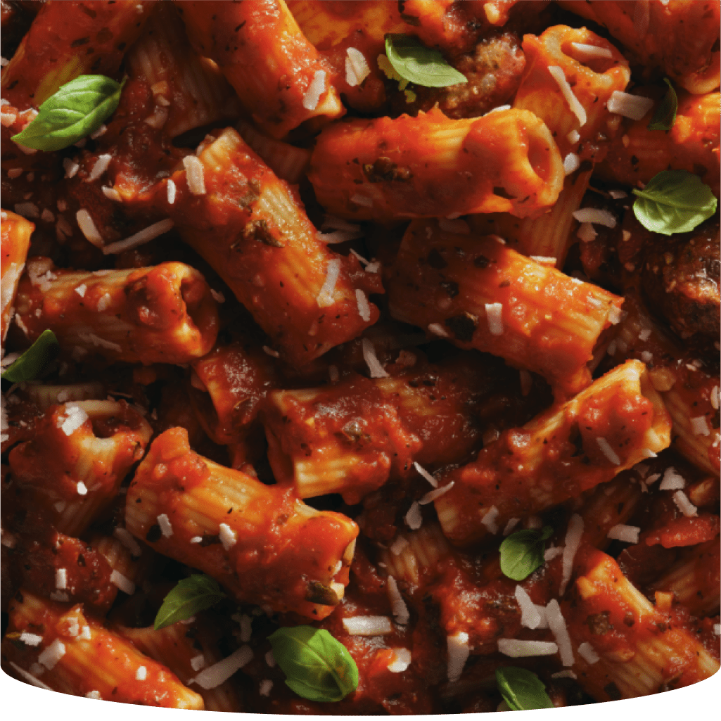 Close-up of rigatoni pasta with Classico tomato sauce, meatballs, and grated cheese, garnished with fresh basil leaves.