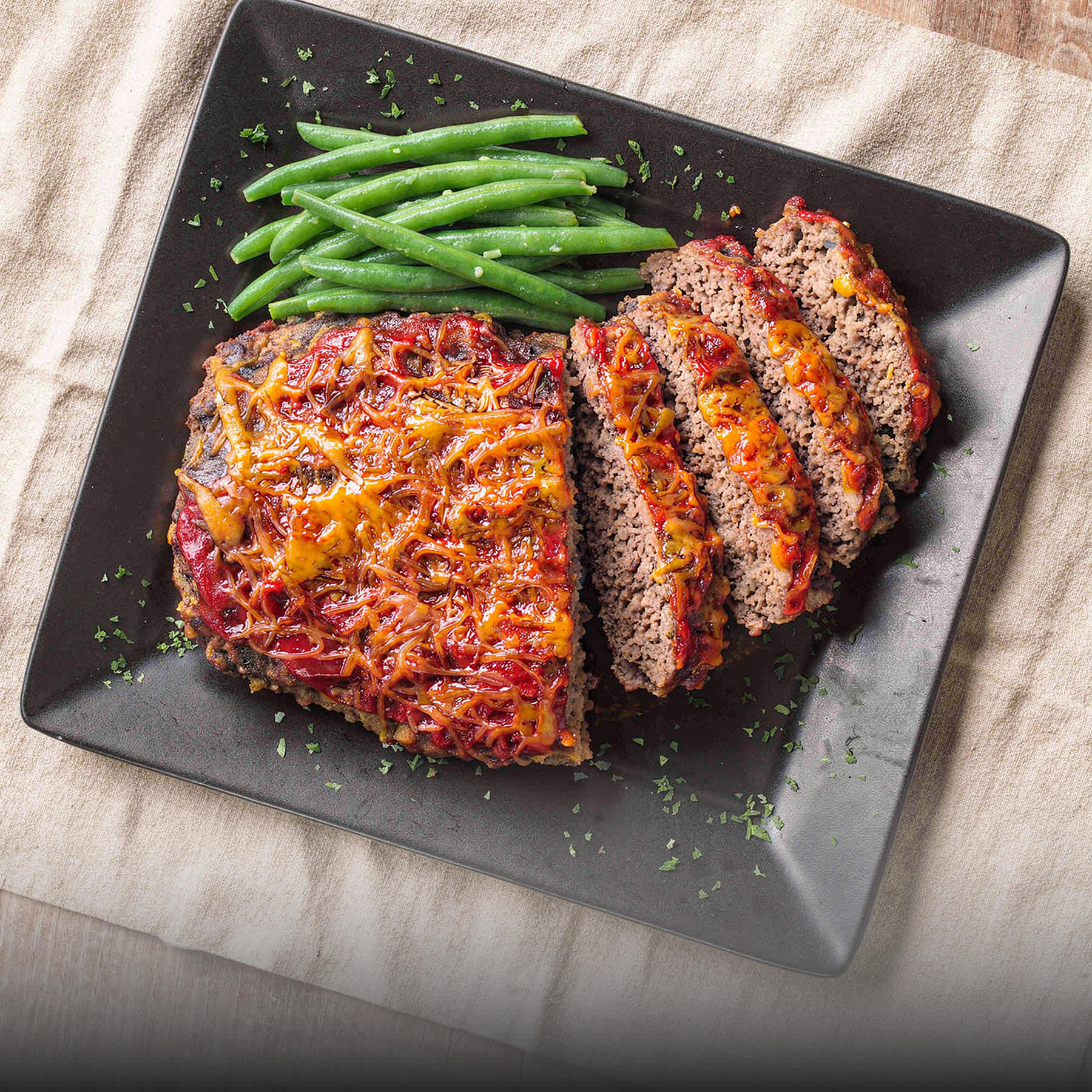 Sliced meatloaf topped with melted cheese and red glaze, served with fresh green beans on a dark square plate.