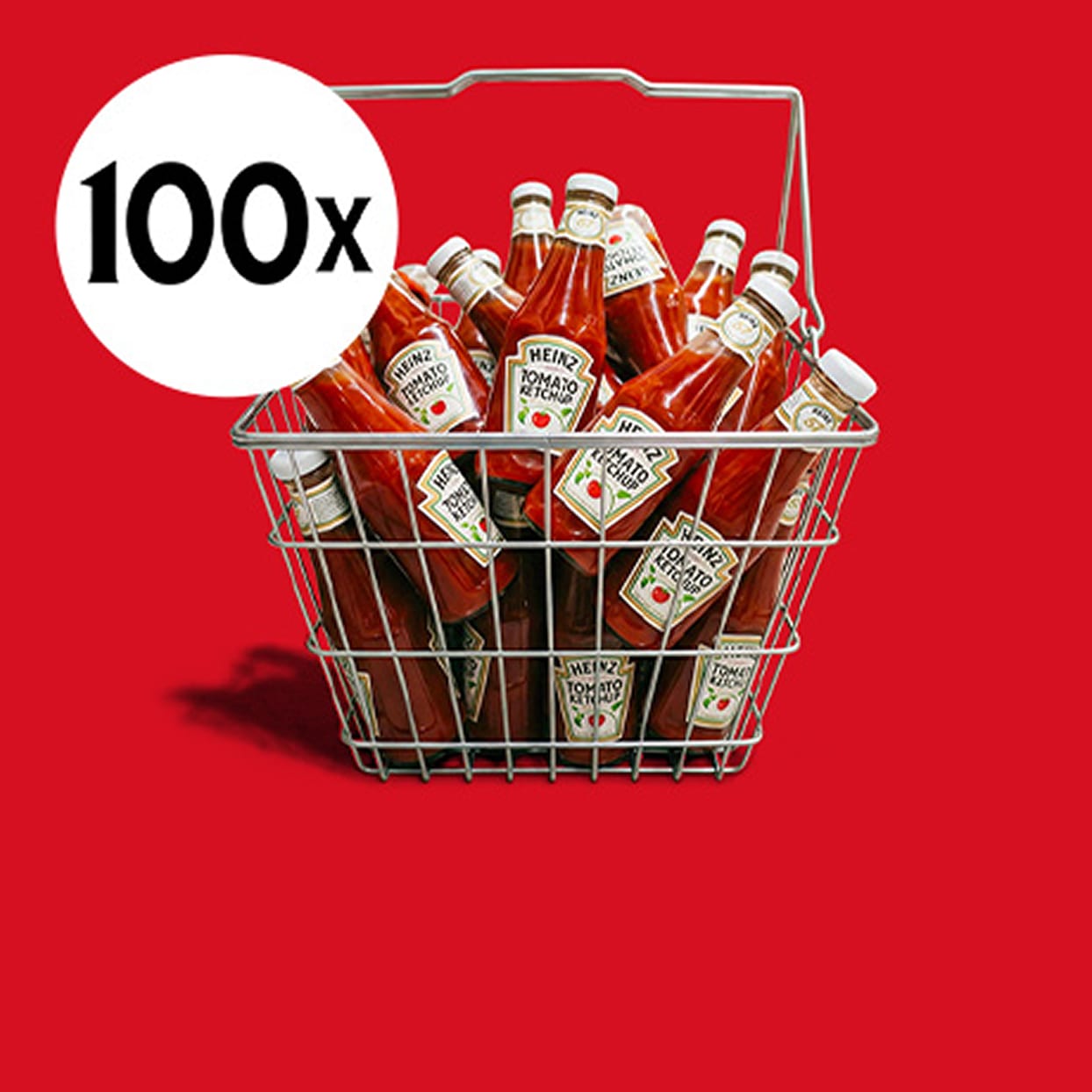 Metalowy koszyk zakupowy pełen butelek ketchupu Heinz z białą grafiką „100x” na czerwonym tle