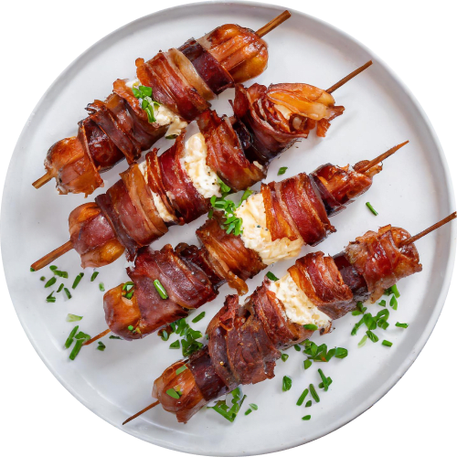 Overhead shot of four sausage and cheese skewers arranged on a white plate. Each skewer features cooked sausage pieces alternating with chunks of cheese, all wrapped in crispy bacon. The plate is garnished with chopped chives.
