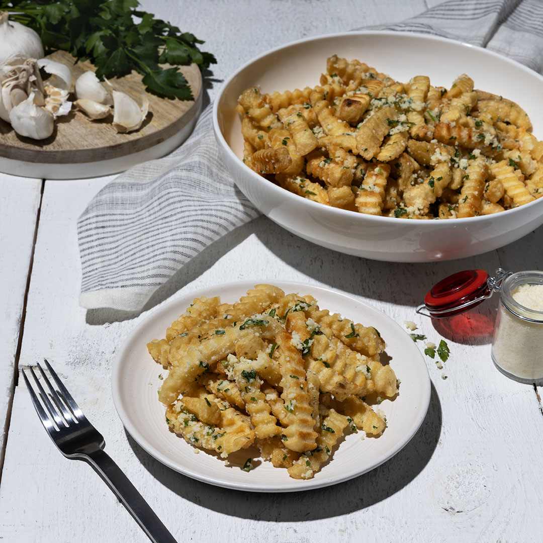 Crispy golden fries seasoned with fresh garlic, parmesan cheese, and parsley. A plate of the fries sits in the foreground with a fork, and a larger bowl of fries is visible in the background, along with fresh ingredients and a napkin, all arranged on a white wooden table.
