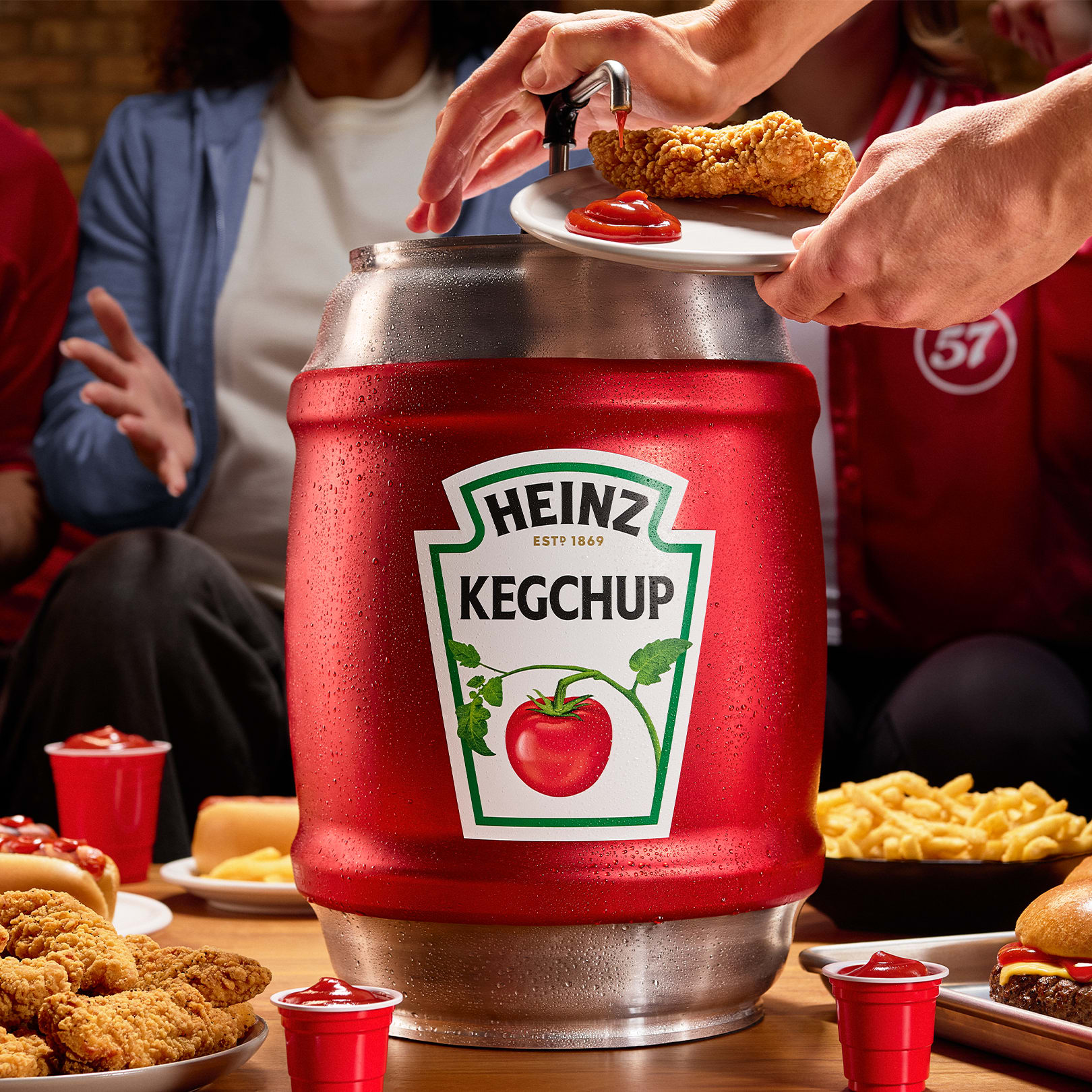 A hand dispensing ketchup from a "Heinz Kegchup" keg onto a plate of chicken tenders, surrounded by burgers and fries.
