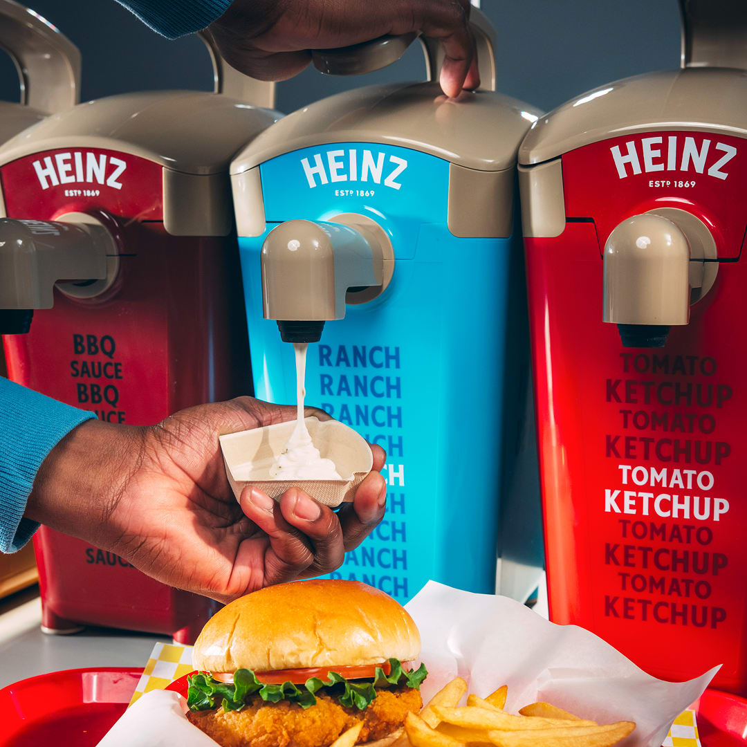 A hand dispensing ranch dressing from a Heinz condiment dispenser, with a burger and fries in the foreground.