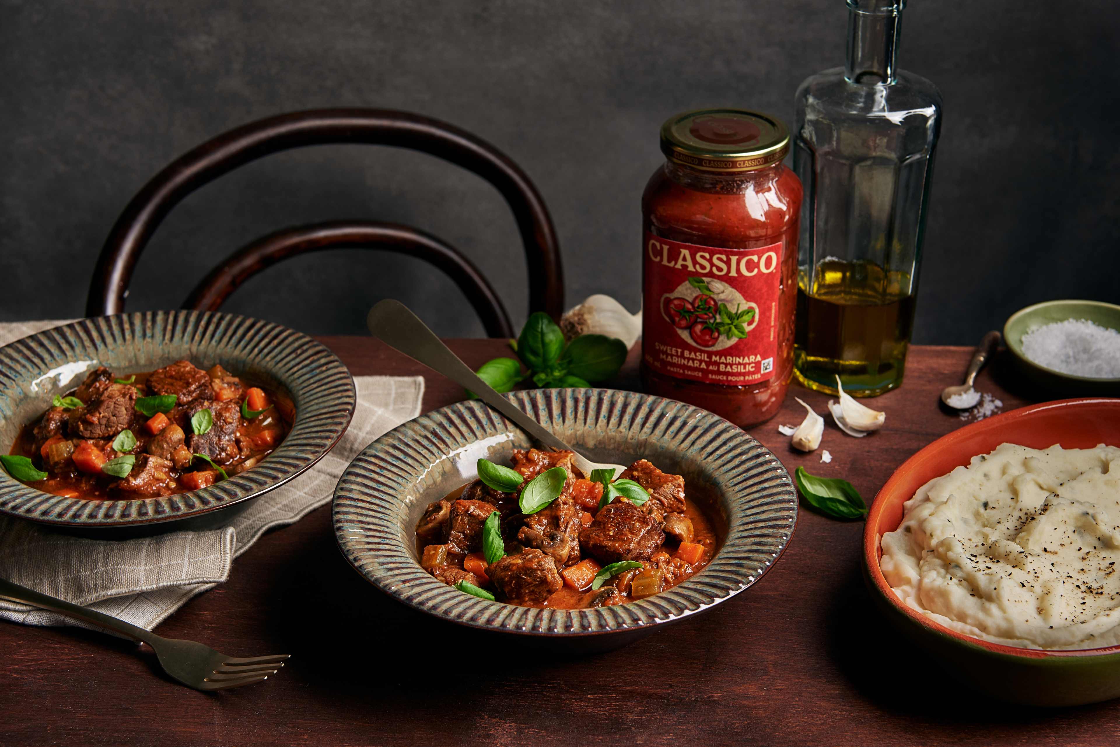 Two bowls of beef stew and vegetable stew, alongside a bowl of mashed potatoes, with fresh basil, garlic, olive oil and salt. A jar of Classico Sweet Basil Marinara sauce sits behind the stew.