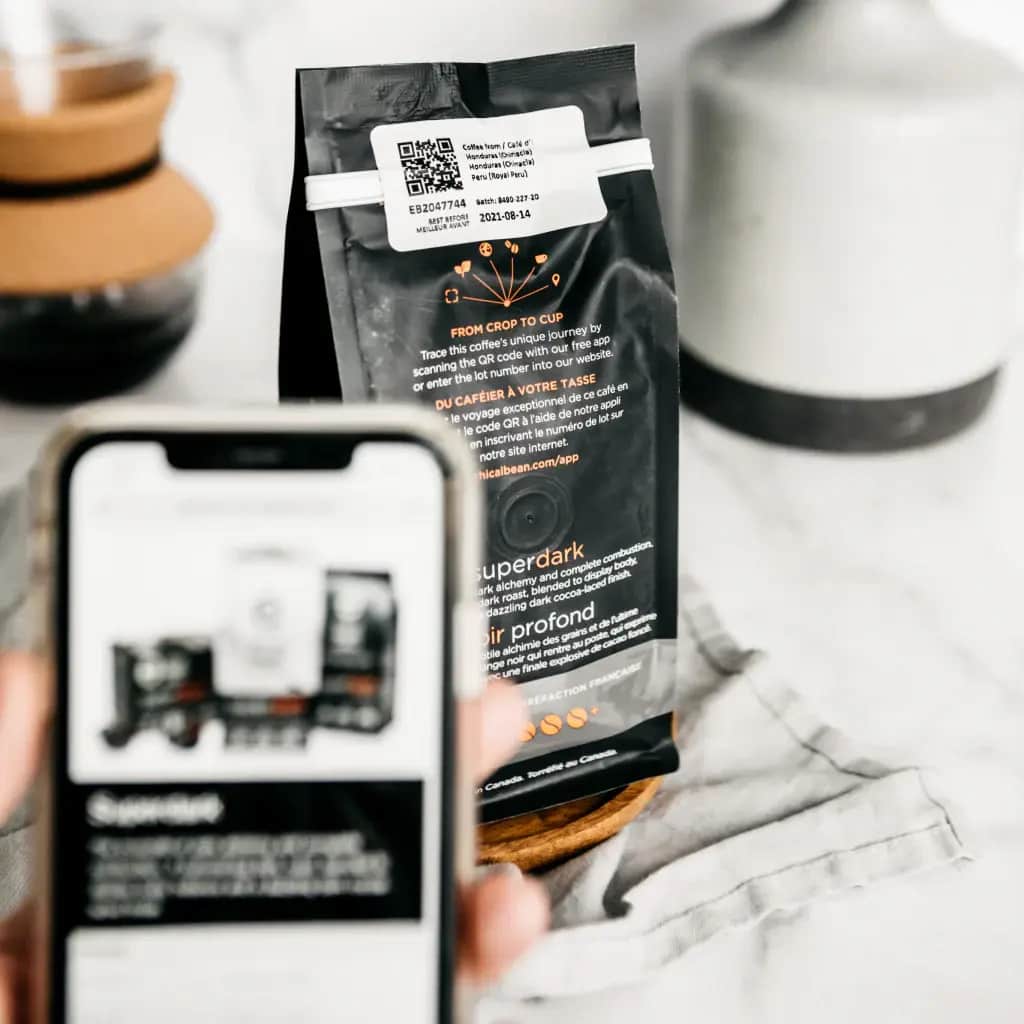 A hand holds a smartphone, scanning a QR code on an Ethical Bean Coffee Superdark bag. The bag sits on a wooden coaster atop 