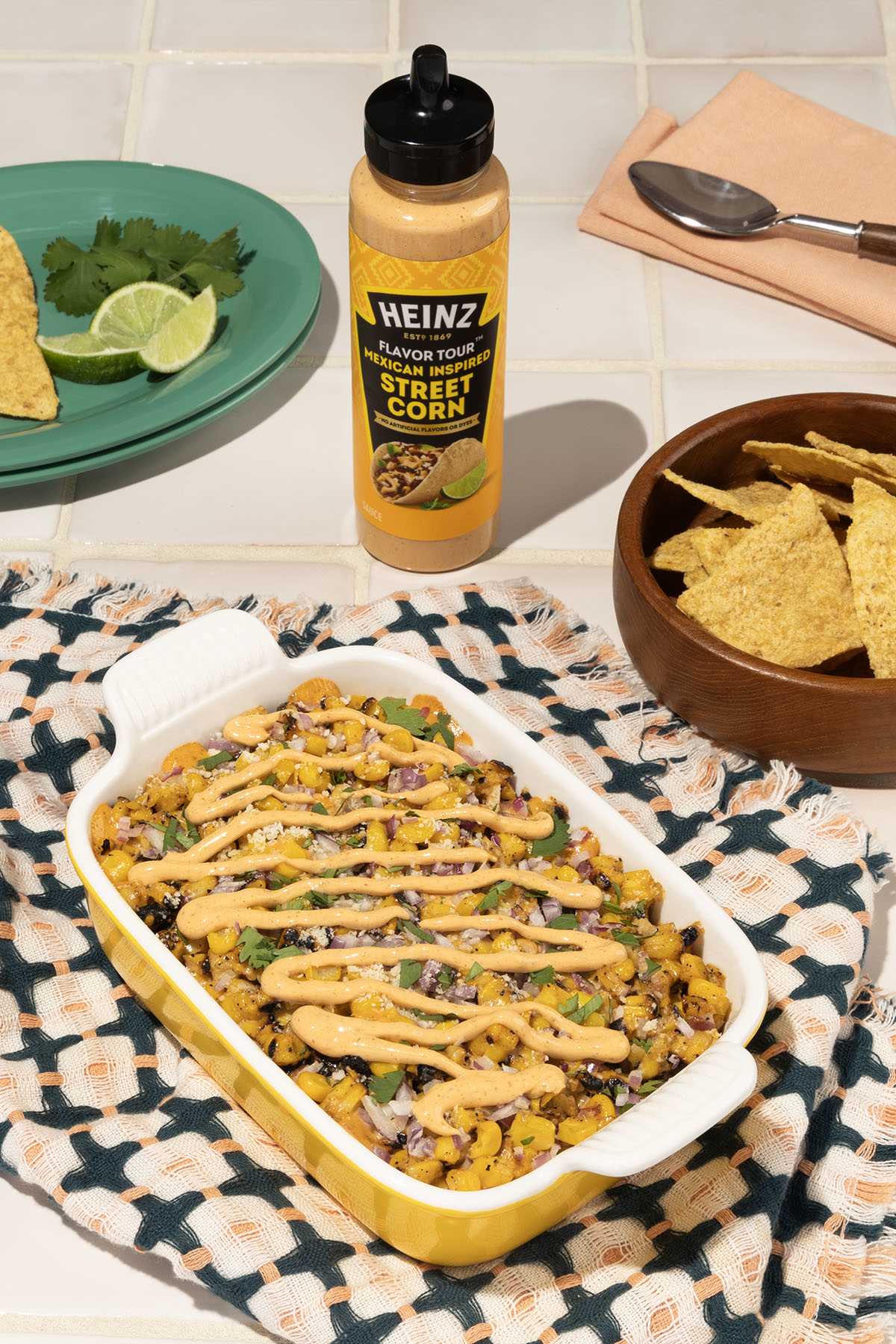 A yellow baking dish filled with Mexican street corn dip is placed on a patterned cloth. The dip is topped with a creamy, ora