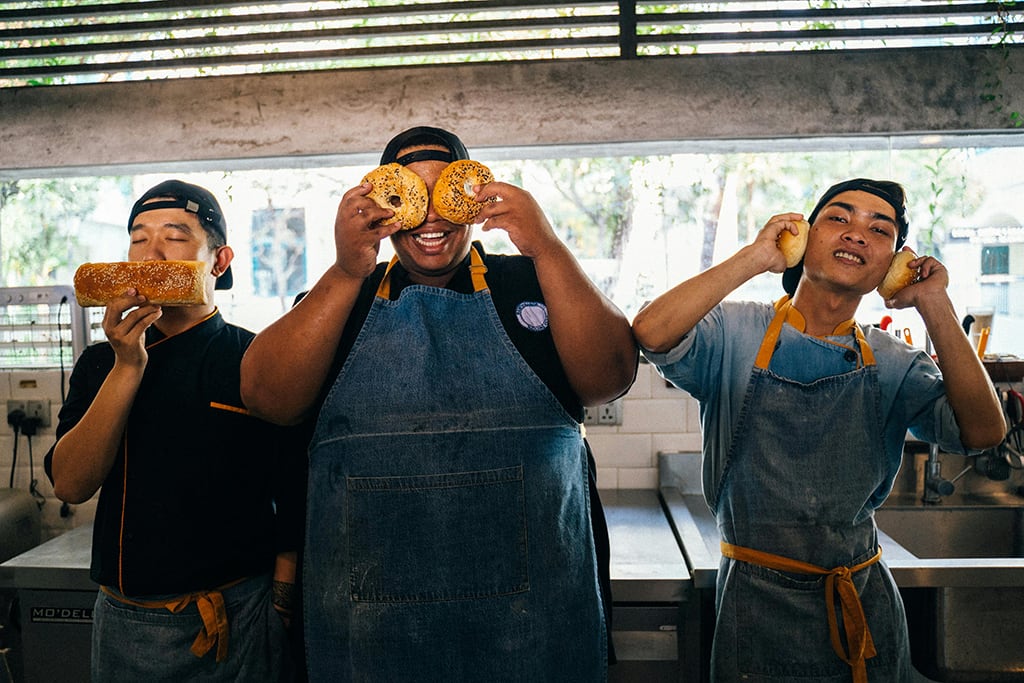 Three playful chefs in a kitchen, one smelling bread, one with bagels on eyes, and one with rolls on ears, all smiling.