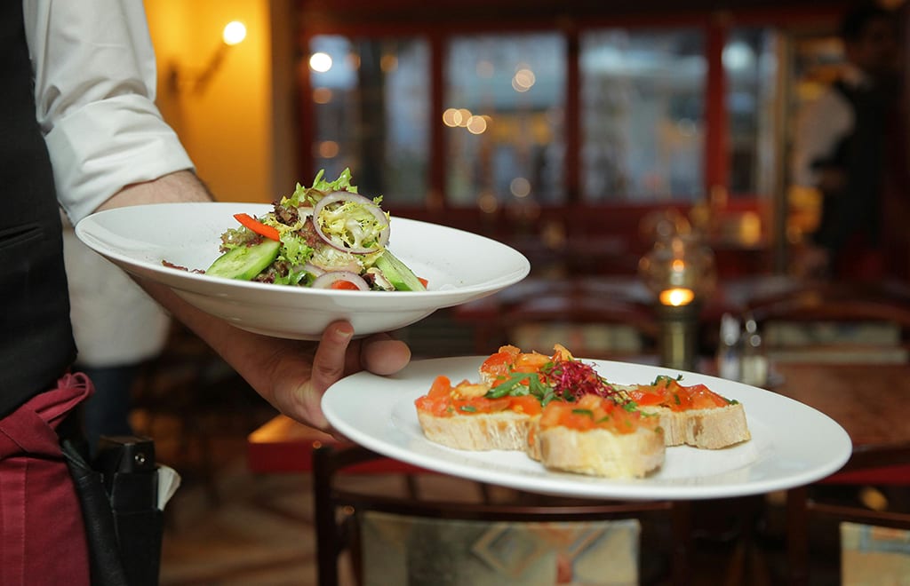 A waiter holds two plates of food: a fresh salad and bruschetta, in a softly lit restaurant setting.