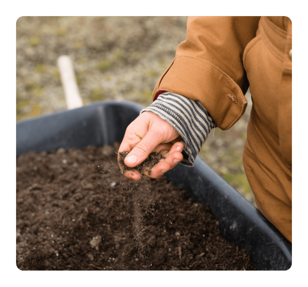 A close-up view of a person's hand, clad in a striped sleeve and brown work jacket, gently sifting dark, rich soil. The soil 