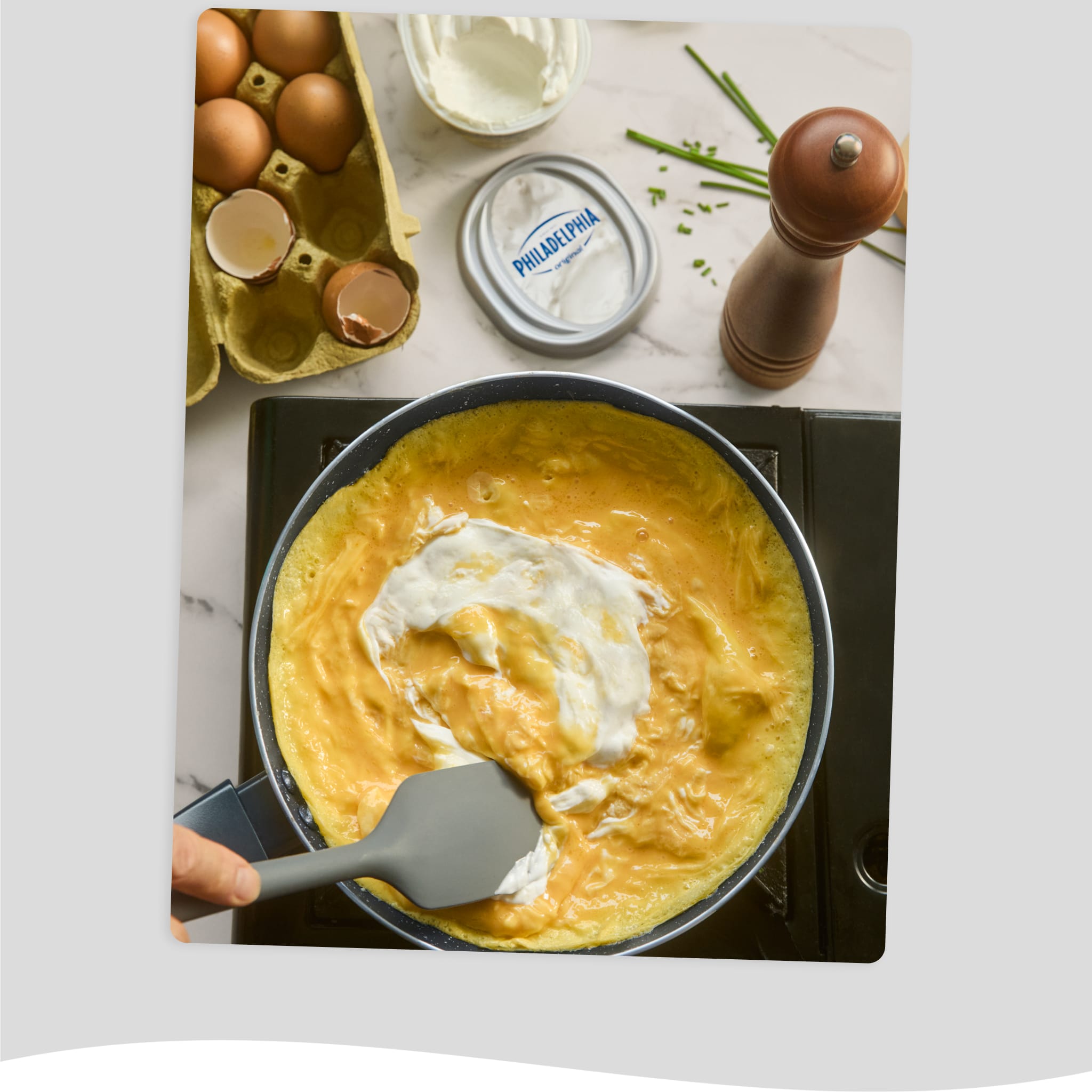 Scrambling eggs in a pan with Philadelphia Cream Cheese, surrounded by fresh eggs and chives.