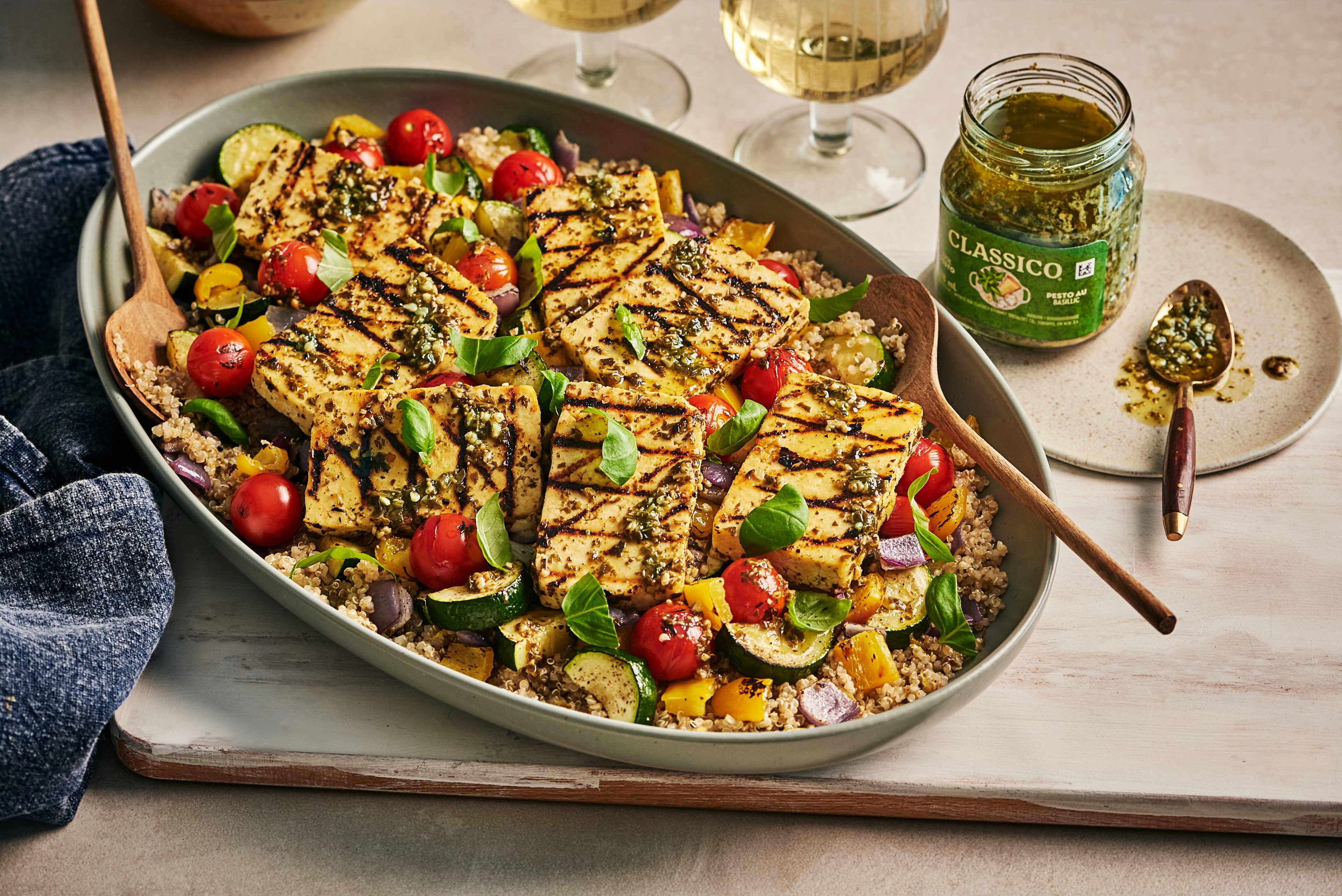 Un plat de tranches de tofu grillées servies sur un lit de quinoa et de légumes variés. Un pot de pesto Classico et une cuillerée de pesto se trouvent à côté du plat. Deux verres de vin blanc sont à l'arrière-plan.