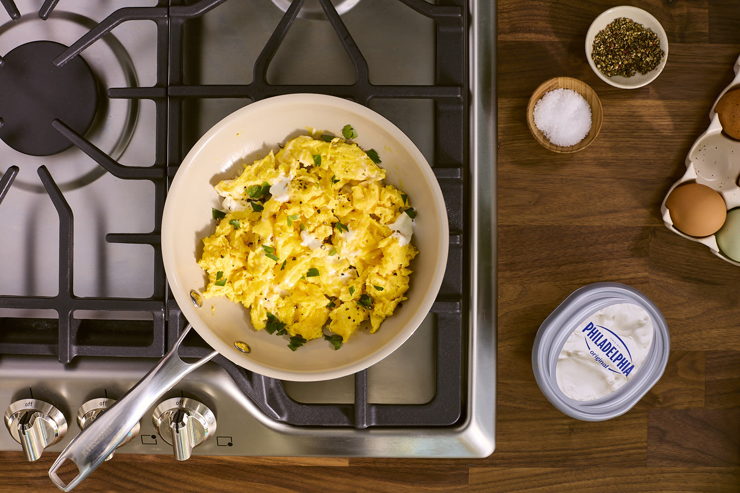 A pan of creamy scrambled eggs sits on a stovetop. Ingredients including Philadelphia cream cheese, eggs, salt, pepper, and parsley are arranged on the wooden countertop beside the stove.