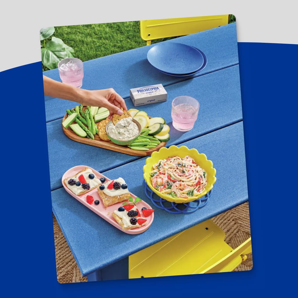A vibrant outdoor picnic scene featuring a spread of delicious treats. A hand reaches for a Philadelphia cream cheese-topped 