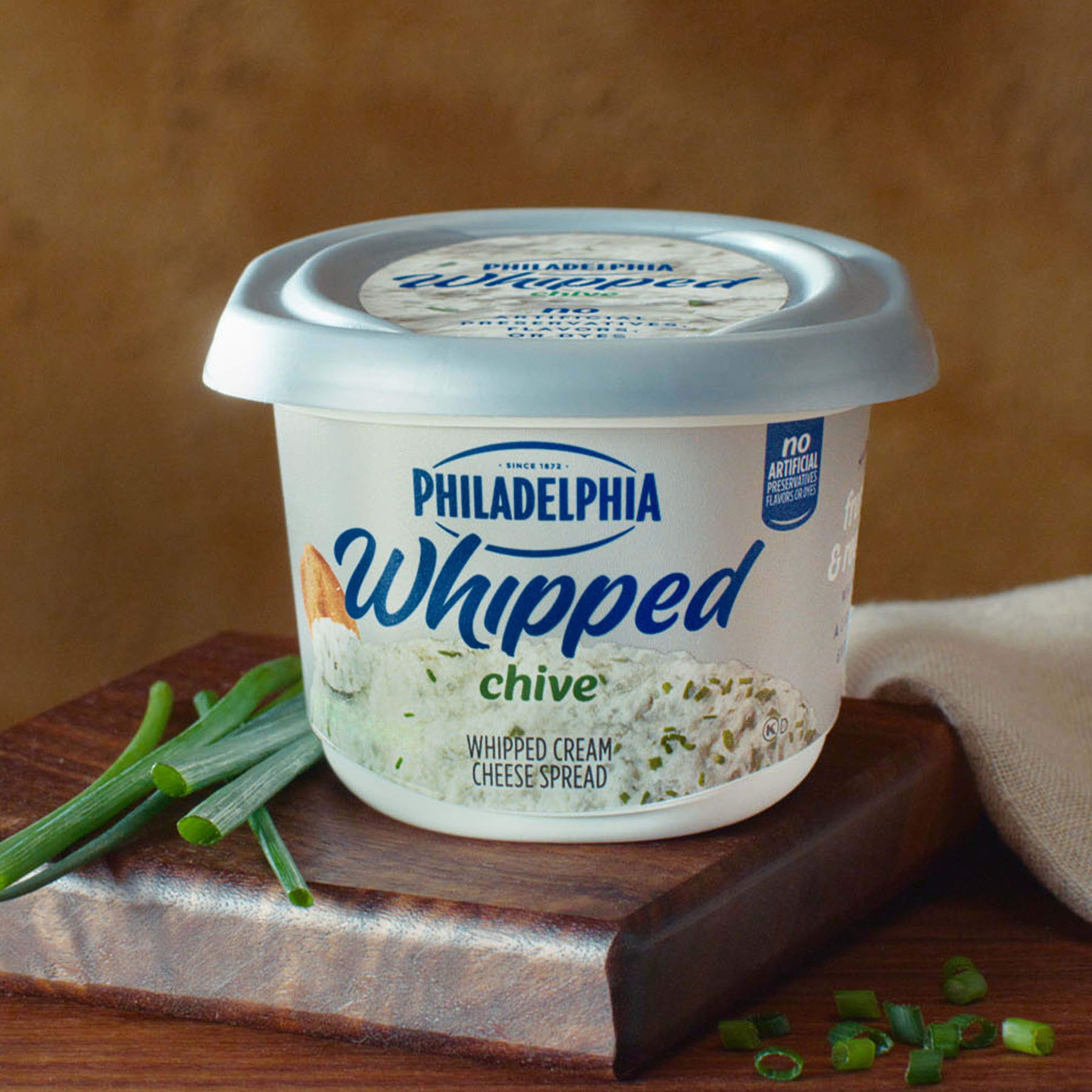 A tub of Philadelphia Whipped Chive cream cheese on a wooden block with fresh green chives.