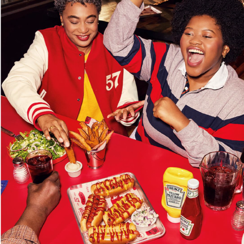 This image represents a filter on the Heinz product hub, showcasing products perfect for entertaining. It features friends enjoying a lively gathering with a platter of hot dogs topped with Heinz condiments, accompanied by sides of sweet potato fries and a fresh salad. The vibrant red and white decor, along with the joyful expressions of the people, creates a sense of fun and togetherness.