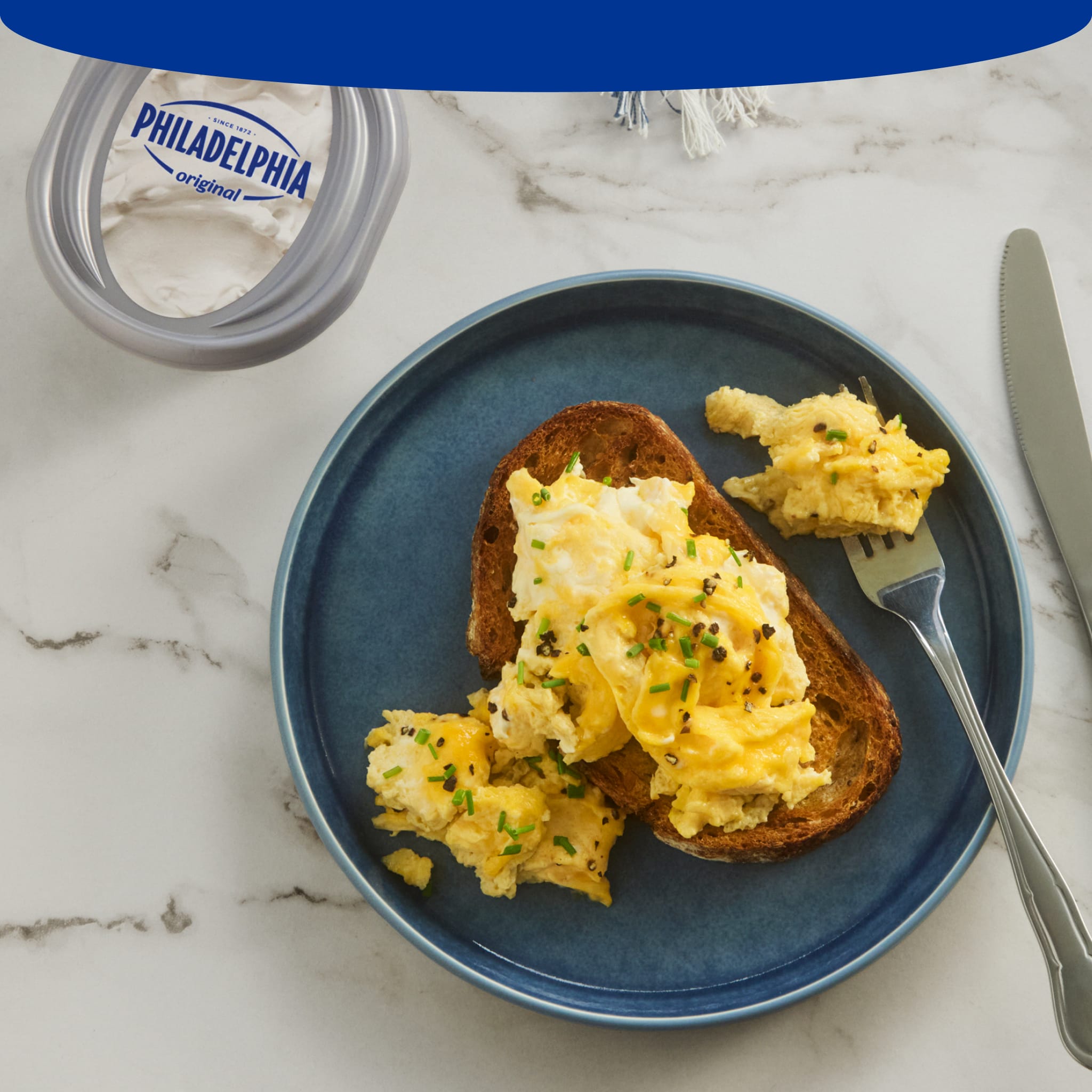 Creamy scrambled eggs with chives on sourdough toast next to a tub of Philadelphia Cream Cheese.