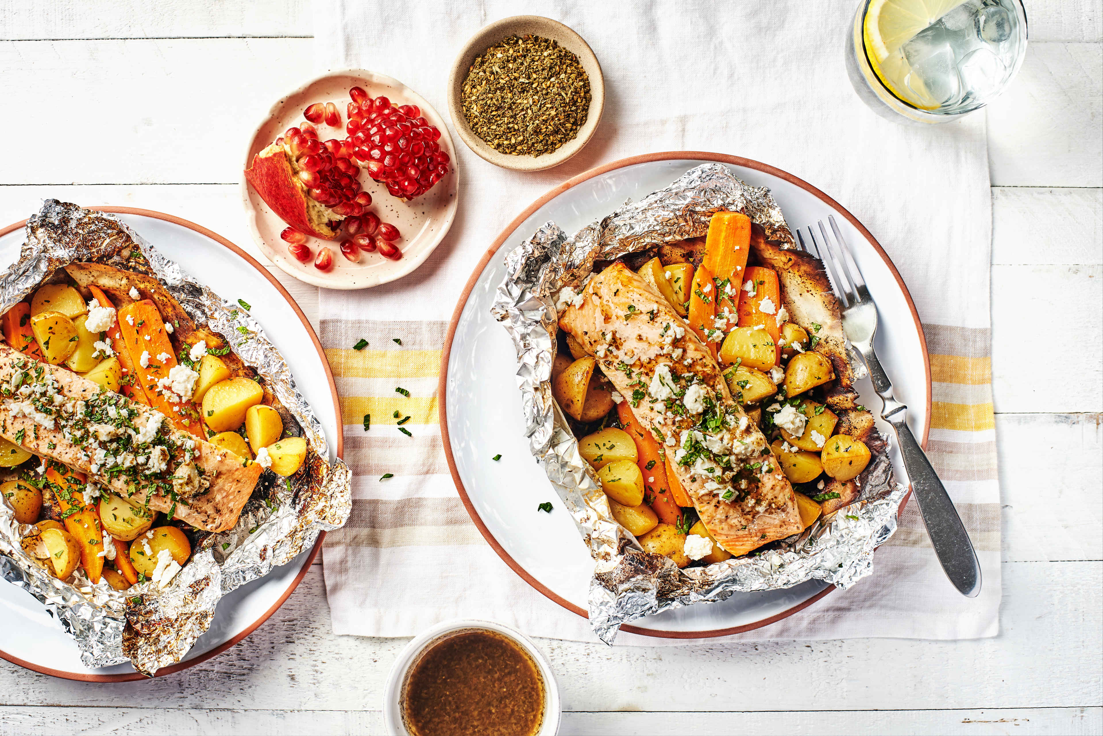 wo servings of Salmon and Vegetable Foil Packets with Za'atar, featuring Pure J.L. KRAFT Pomegranate Za'atar Dressing & Marinade, photographed overhead on white wood table, complete meal, easy recipe, healthy eating.