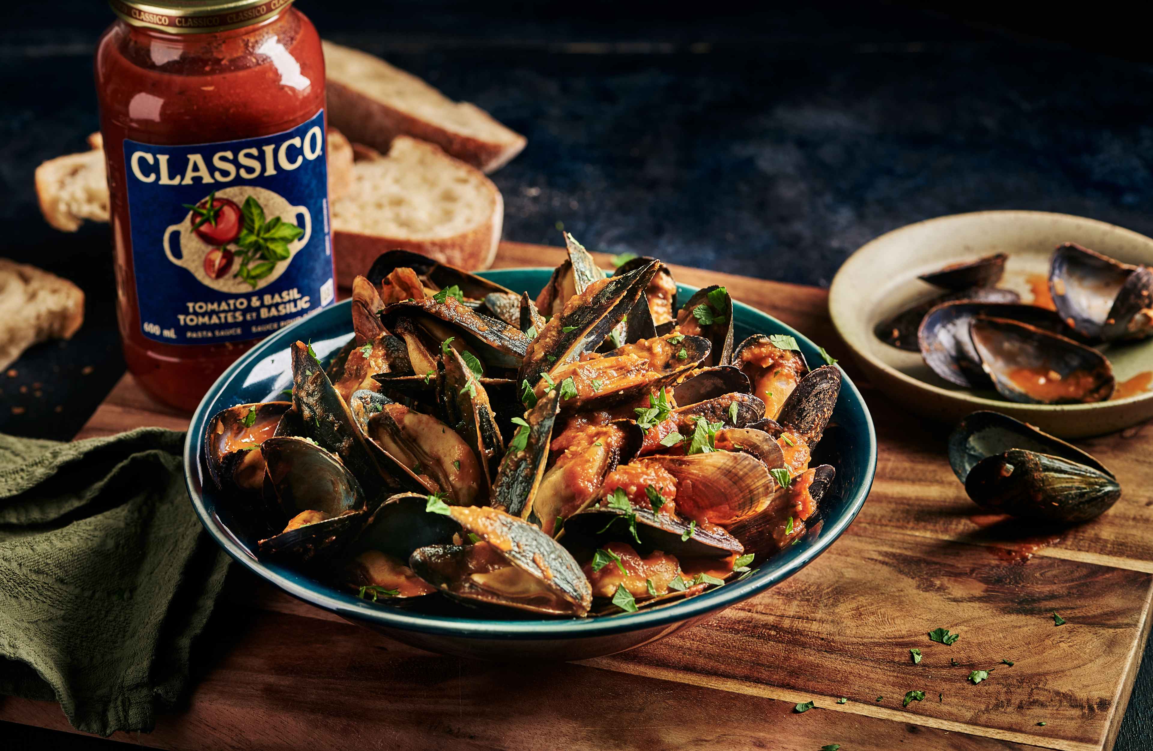 Un gros plan d'un bol de moules à la vapeur, nappées d'une sauce pour pâtes Classico Tomates et Basilic aux couleurs éclatantes. Du persil frais est saupoudré sur le dessus, ajoutant une touche de vert aux riches teintes rouge orangé. Des tranches de pain croustillant sont visibles à l'arrière-plan, à côté du pot de sauce Classico, ce qui suggère un repas savoureux et réconfortant.