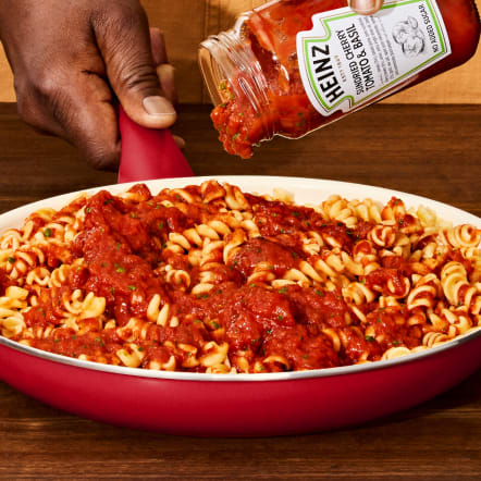 A hand pours Heinz Simmered Cherry Tomato & Basil Sauce from a jar onto rotini pasta in a red pan, set against a warm wood background. This image represents the "Sauce" meal type filter for recipes.