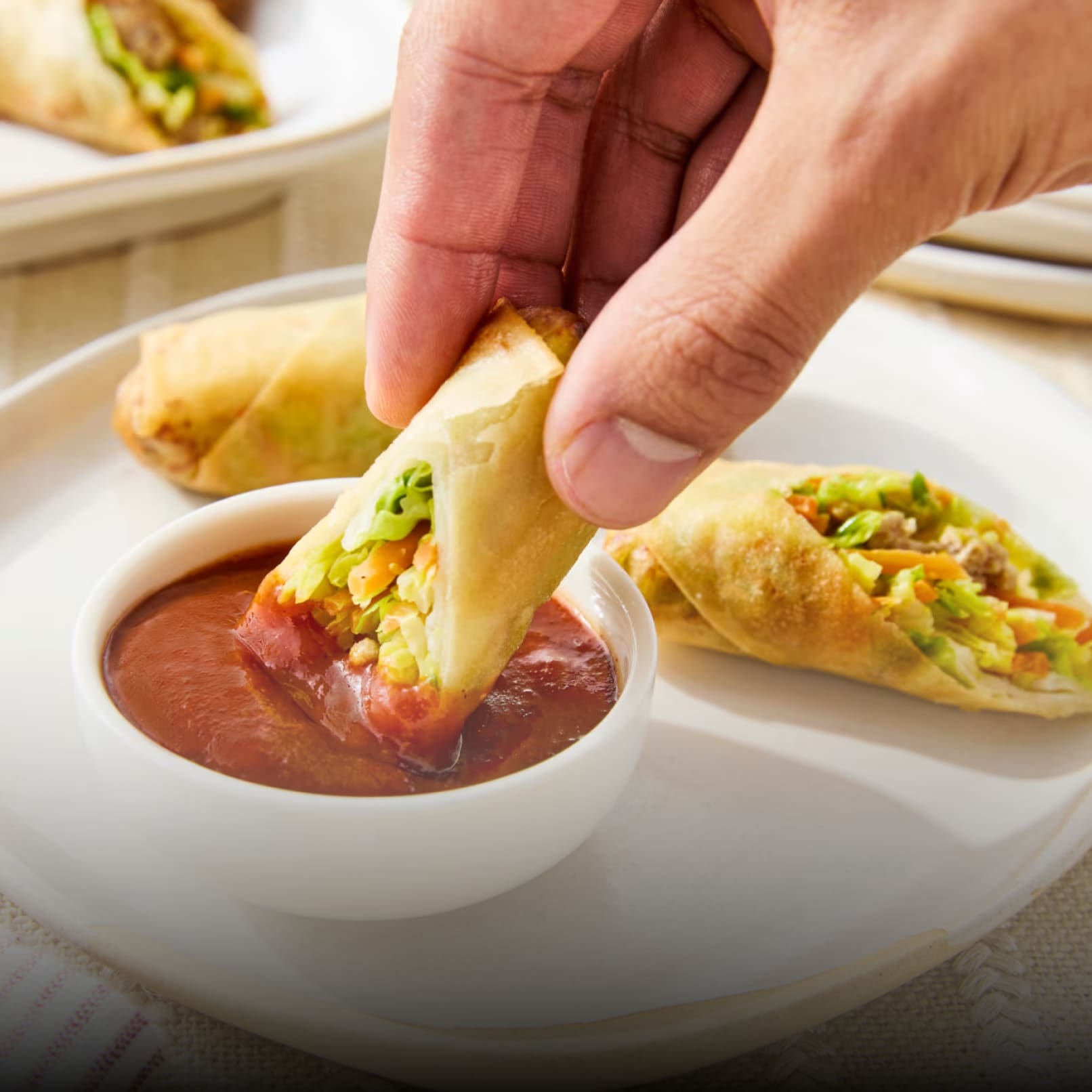 Close-up of a hand dipping a crispy spring roll filled with colorful vegetables into a small white bowl of vibrant, reddish-o
