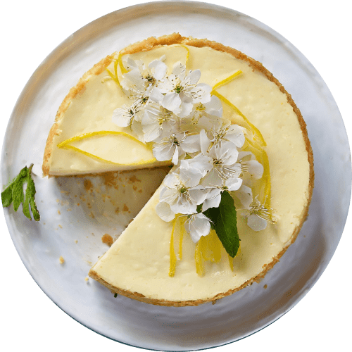 Overhead shot of a whole lemon cheesecake on a white plate, with one slice removed. The cheesecake is decorated with white spring blossoms, thin lemon zest curls, and fresh mint leaves.