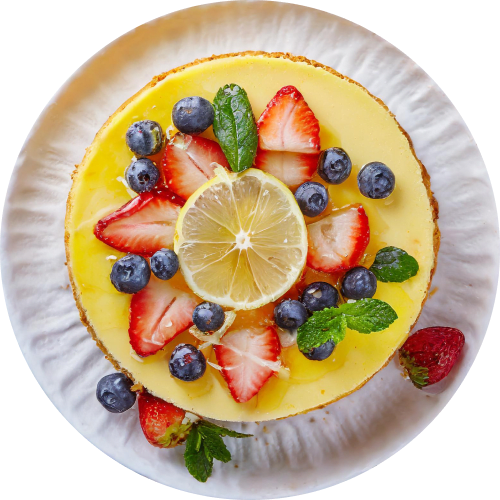 Overhead shot of a whole lemon cheesecake on a white plate. The cheesecake is topped with a thin layer of lemon curd, fresh strawberry slices, blueberries, mint leaves, and a center slice of lemon. A drizzle of honey adds a touch of sweetness.