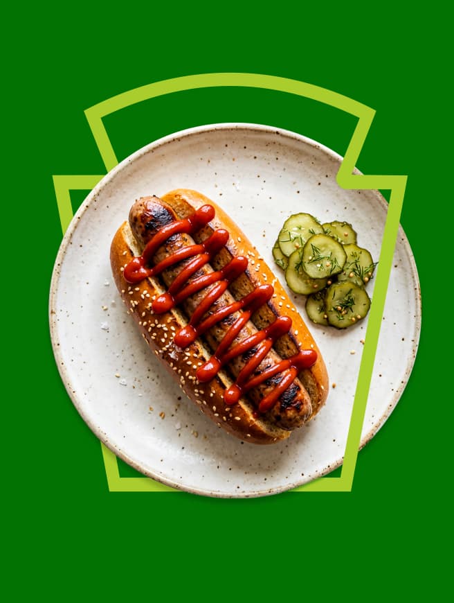  Grilled hot dog with ketchup on a sesame bun and pickles on a plate, framed by a green Heinz keystone on a green background.