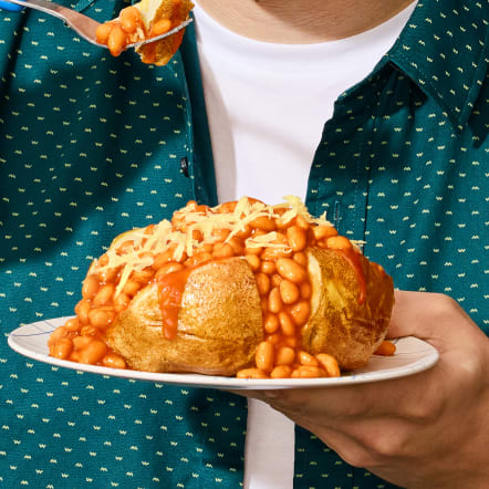 A person wearing a teal patterned shirt holds a plate with a loaded baked potato. The potato is topped with baked beans in tomato sauce and grated cheese. A fork lifts a bite of beans, emphasizing the meal's appeal. This image is for a "Dinner" meal type filter.