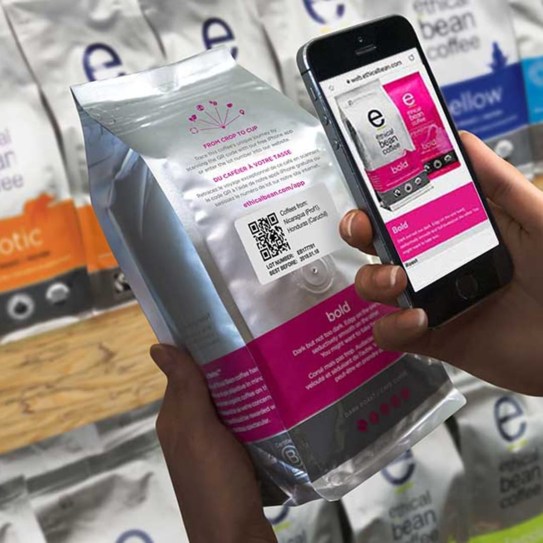 A person uses a smartphone to scan a QR code on an Ethical Bean Coffee bag. The bag, held in another hand, features pink and 