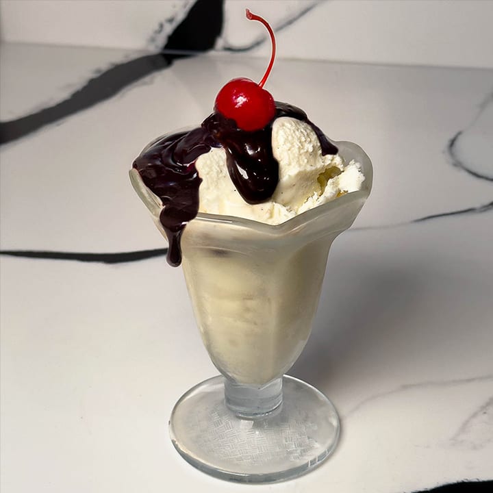 A decadent dessert of vanilla ice cream topped with rich, dripping Velveeta chocolate fudge sauce and a bright red cherry.