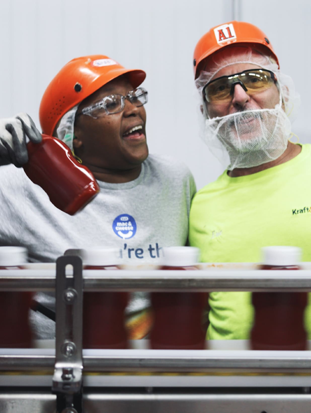 Two Kraft Heinz employees, wearing safety helmets and protective eyewear, smile as they monitor a production line of ketchup 
