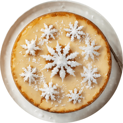 Overhead shot of a whole eggnog cheesecake on a white plate. The cheesecake is decorated with white snowflake candies and a sprinkle of grated nutmeg, creating a festive winter theme. A serving spoon is placed next to the cake
