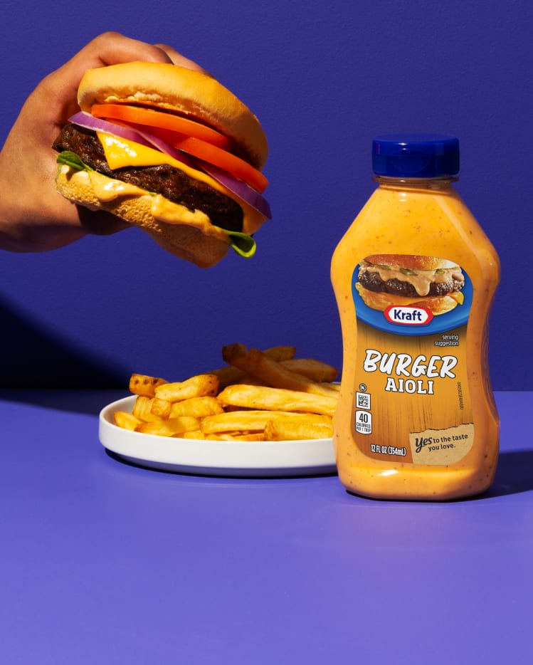 Kraft Burger Aioli bottle next to a burger and fries on a vibrant purple background.