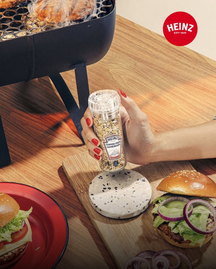 A hand holding a Heinz burger seasoning grinder over finished burgers and a grill.