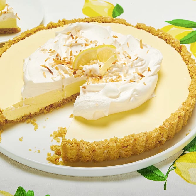 Lemon tart with whipped cream and toasted coconut on a lemon-patterned tablecloth.
