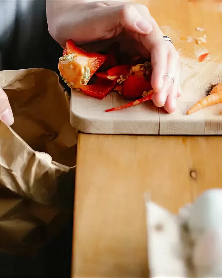 A hand holding discarded red bell pepper scraps over a wooden cutting board, with a paper bag nearby.